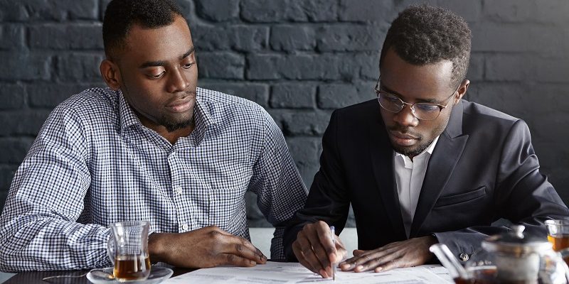 Two confident African-American businessmen dressed formally having serious concentrated looks while signing contract during business meeting at cafe, man with pen showing his partner where to sign